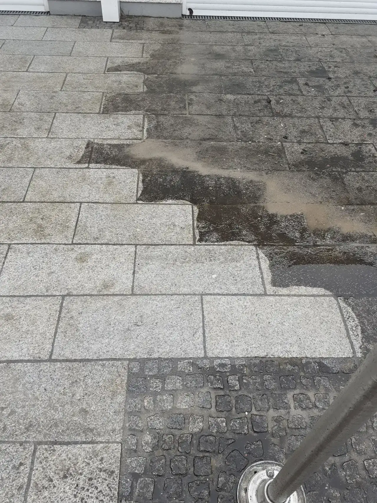 Helle Plattenfläche mit einem dunkel verfärbten Bereich im Hintergrund und einer scharfen Kante dazwischen, unten ein Band aus dunklem Kleinpflaster und ein Geländerpfosten