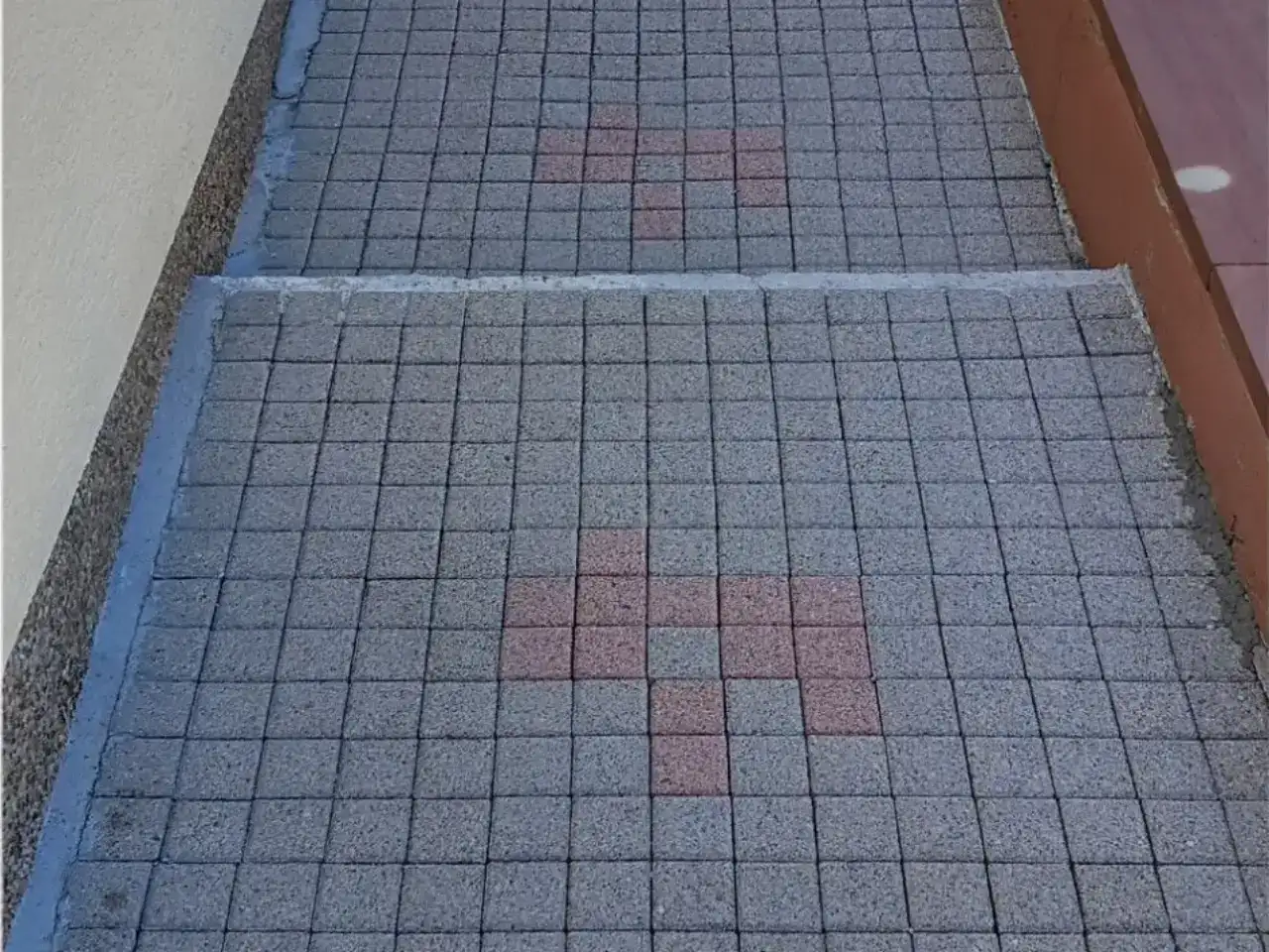 Nachher: Hauszugang aus kleinformatigem Betonpflaster zwischen einer hellen Putzwand und einer Klinkermauer, mit einer Stufe, im Belag zeichnet sich ein rotes Kreuzmuster ab