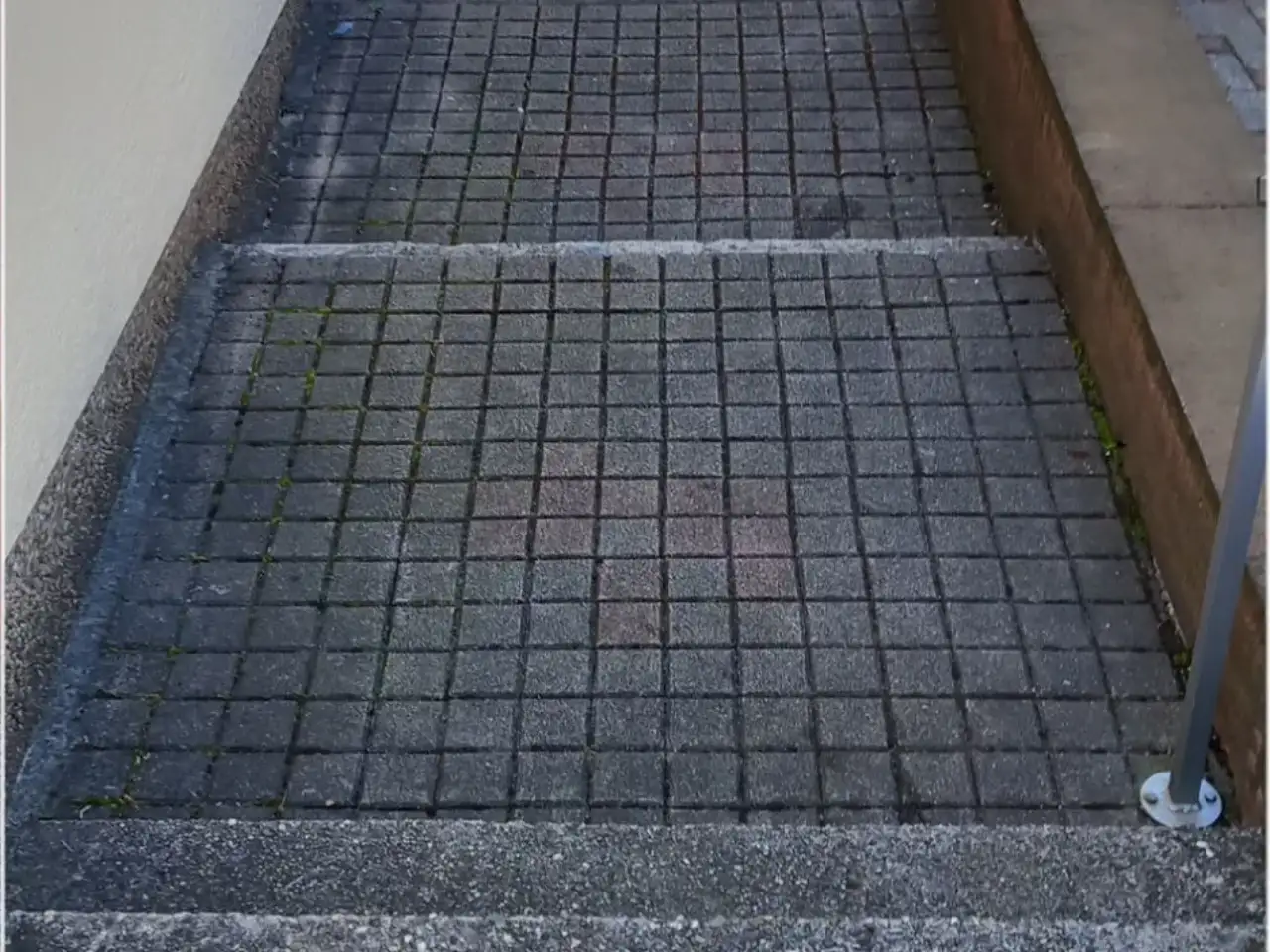 Vorher: Tiefer liegender Hauszugang aus kleinformatigem Betonpflaster zwischen einer hellen Putzwand und einer Klinkermauer, mit einer Stufe und einem Metallpfosten, in den Fugen wächst Moos
