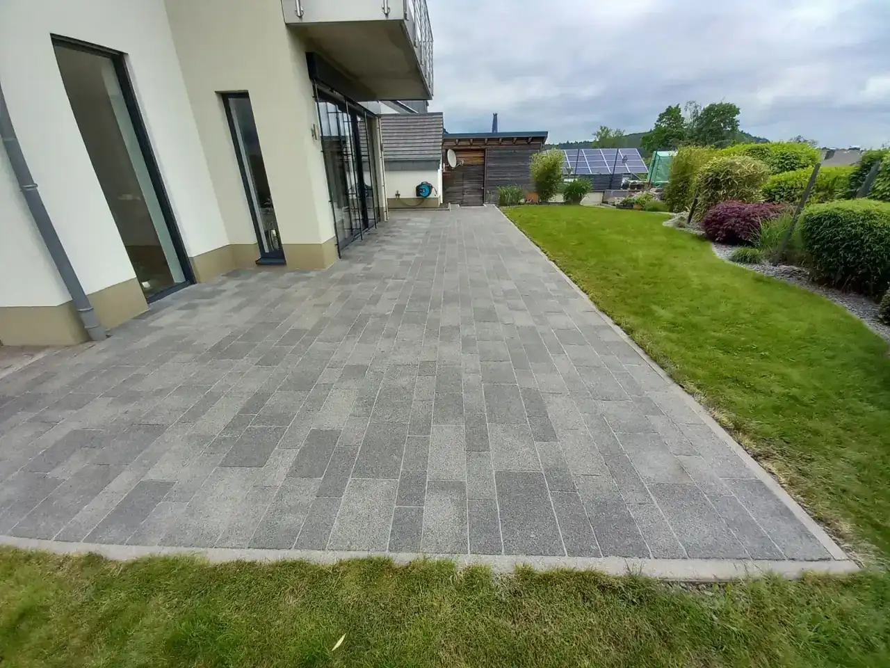 Breiter Plattenbelag in Grautönen entlang einer modernen Hausfassade mit bodentiefen Fenstern, gleichmäßig und ohne Flecken, beidseitig Rasen
