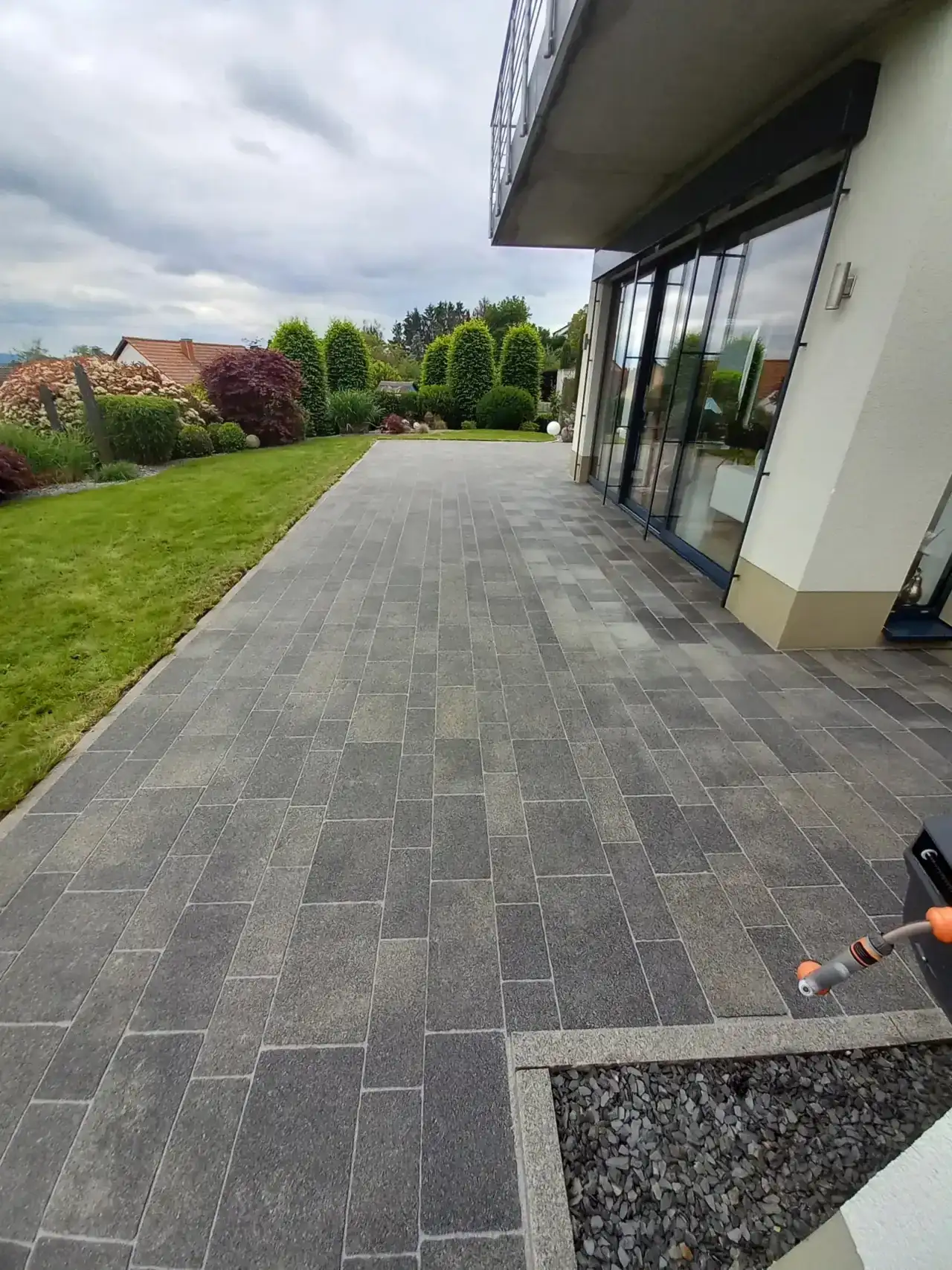 Hausumgang aus grauen Platten entlang einer Fensterfront, gleichmäßig hell, rechts unten liegt die Düse eines Reinigungsgeräts, davor ein Kiesstreifen