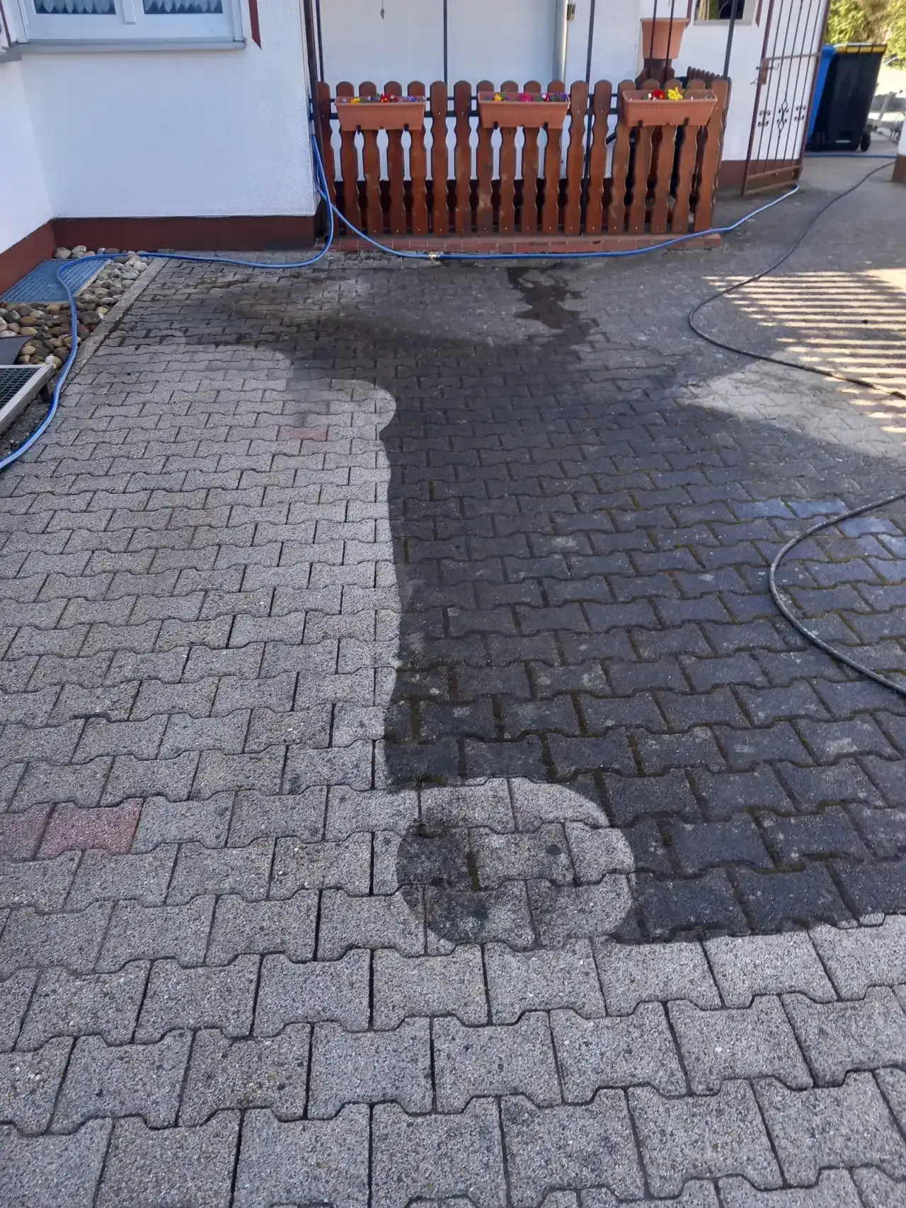 Hoffläche aus Verbundpflaster mit einem hellen Bereich in der Mitte und dunklerem Belag ringsum, ein Schlauch liegt darauf, im Hintergrund ein Holzzaun und eine Hauswand