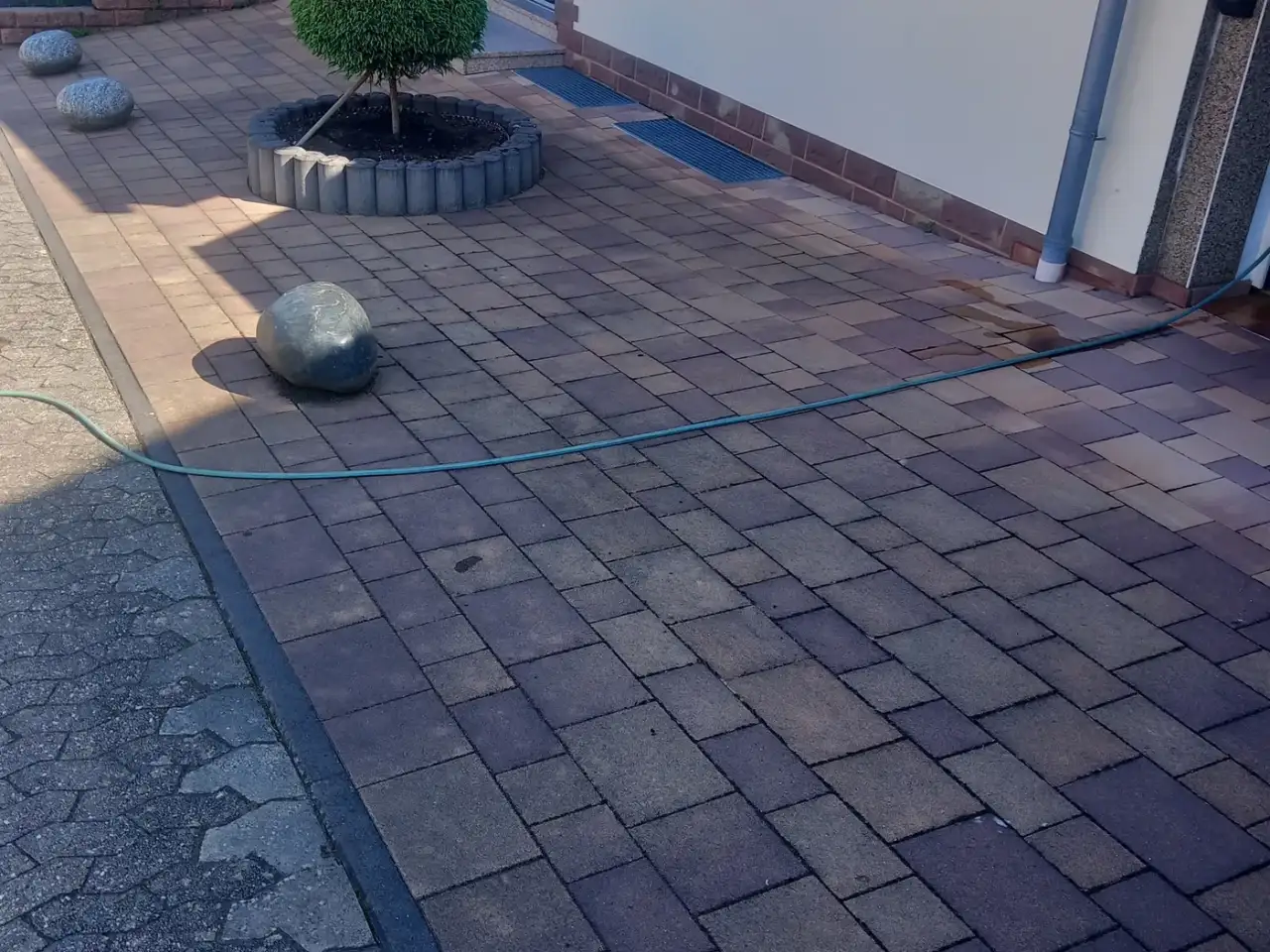 Vorher: Hoffläche aus grauem Betonpflaster am Hauseingang mit einem runden Pflanzbeet und einer Baumkugel, daneben eine Steinkugel und ein Gartenschlauch, der Belag ist stumpf und vergraut