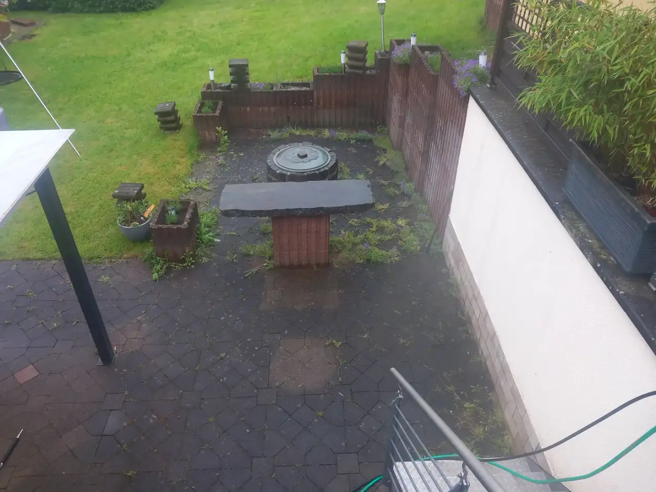 Vorher: Von oben aufgenommener Sitzplatz aus dunklem Betonpflaster mit rundem Brunnendeckel und einem Steintisch, zwischen den Steinen wachsen Kräuter und Gras, ringsum stehen Holzhochbeete und Pflanzkübel