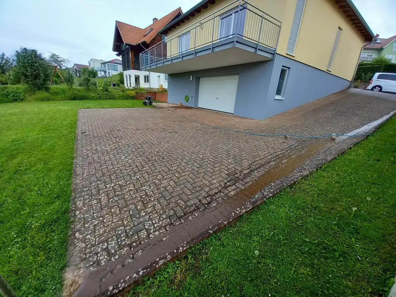 Vorher: Große Stellfläche aus kleinformatigem Betonpflaster vor einem Wohnhaus mit weißem Garagentor, ringsum Rasen, der nasse Belag ist gleichmäßig graubraun verfärbt
